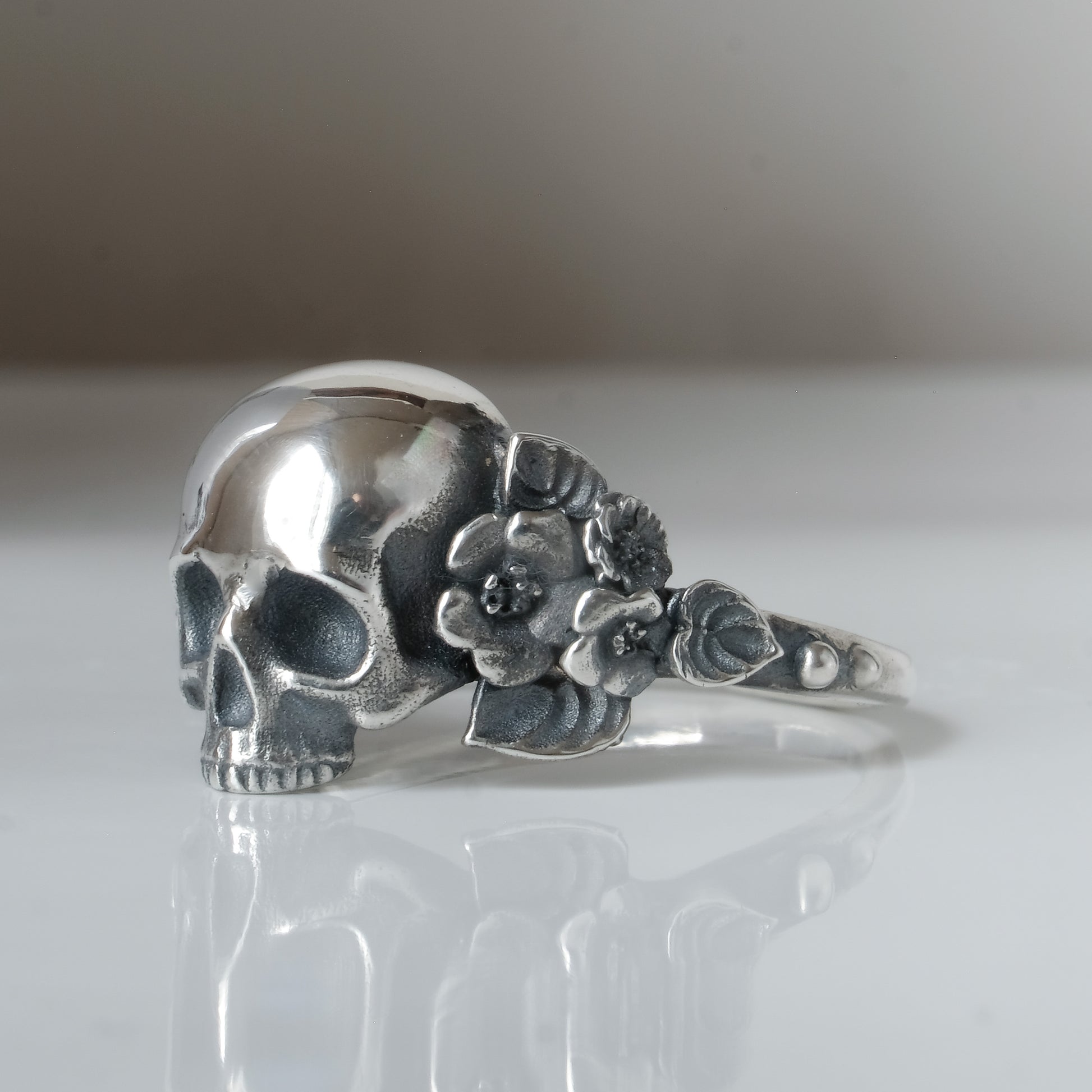 Sterling silver ring with central large skull and flower and leaves cascading down band. Handmade in London by Charlie Shires