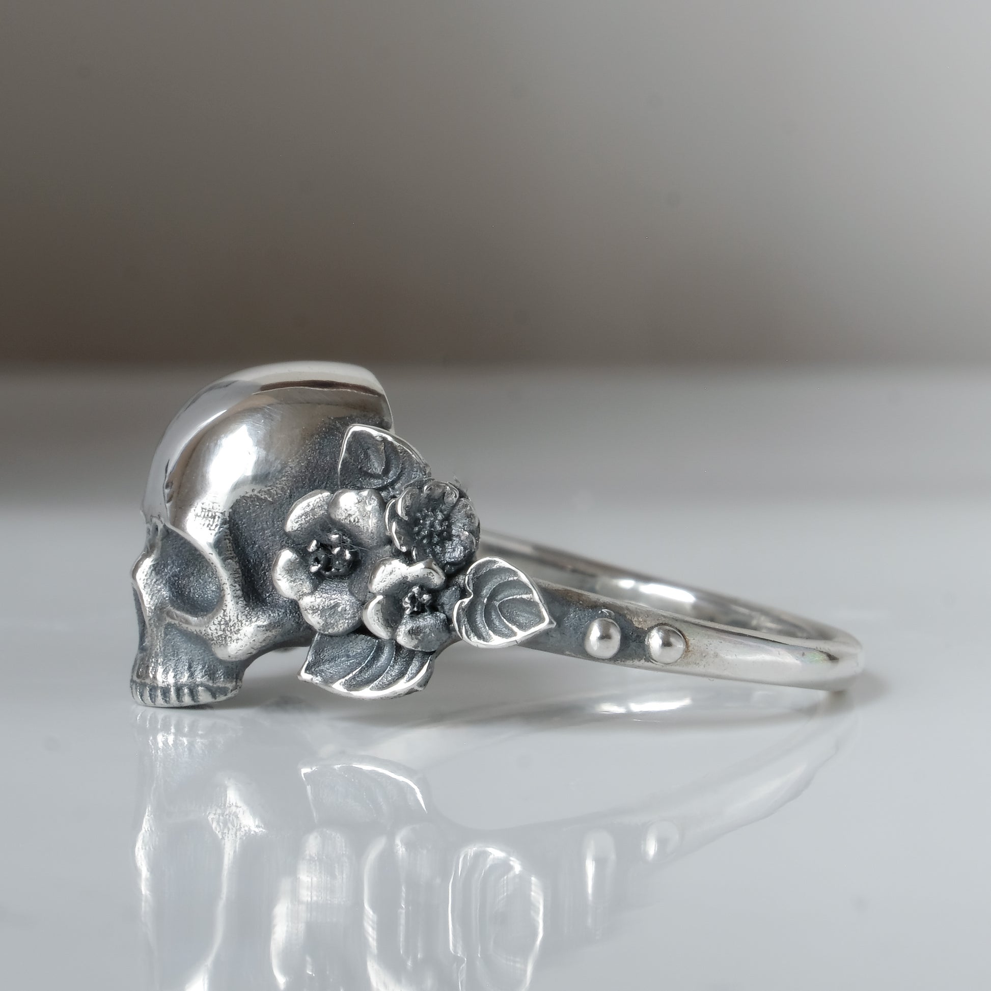 Sterling silver ring with central large skull and flower and leaves cascading down band. Handmade in London by Charlie Shires