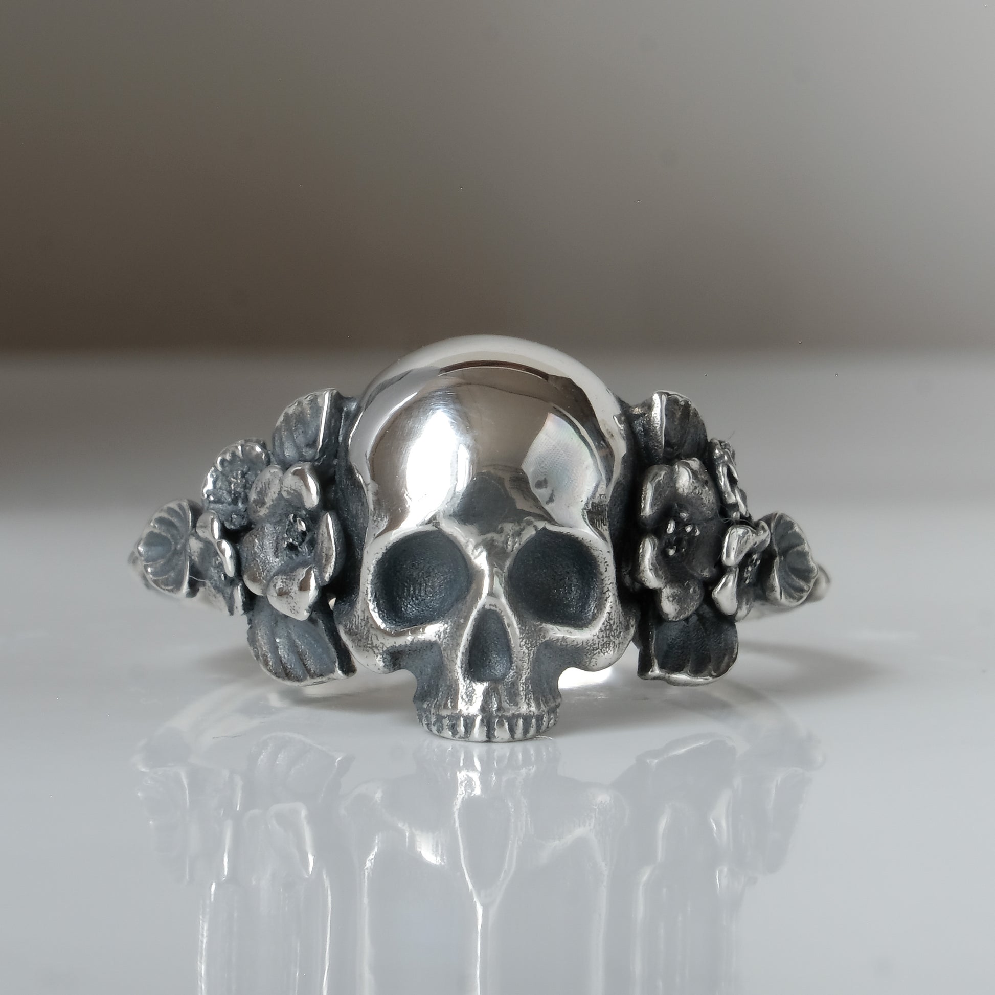 Sterling silver ring with central large skull and flower and leaves cascading down band. Handmade in London by Charlie Shires