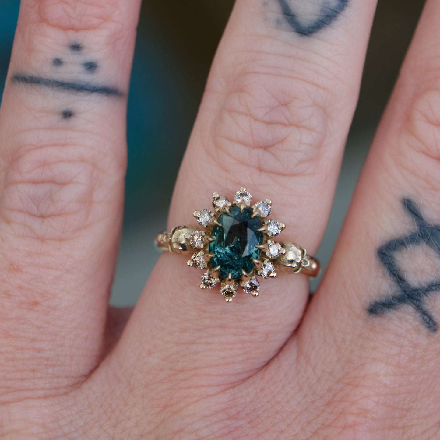Montana sapphire ring with champagne diamonds in a halo/cluster setting, flanked by two skulls. Handmade in 9ct yellow gold on a white background. Handmade in London by Charlie Shires from recycled metals.