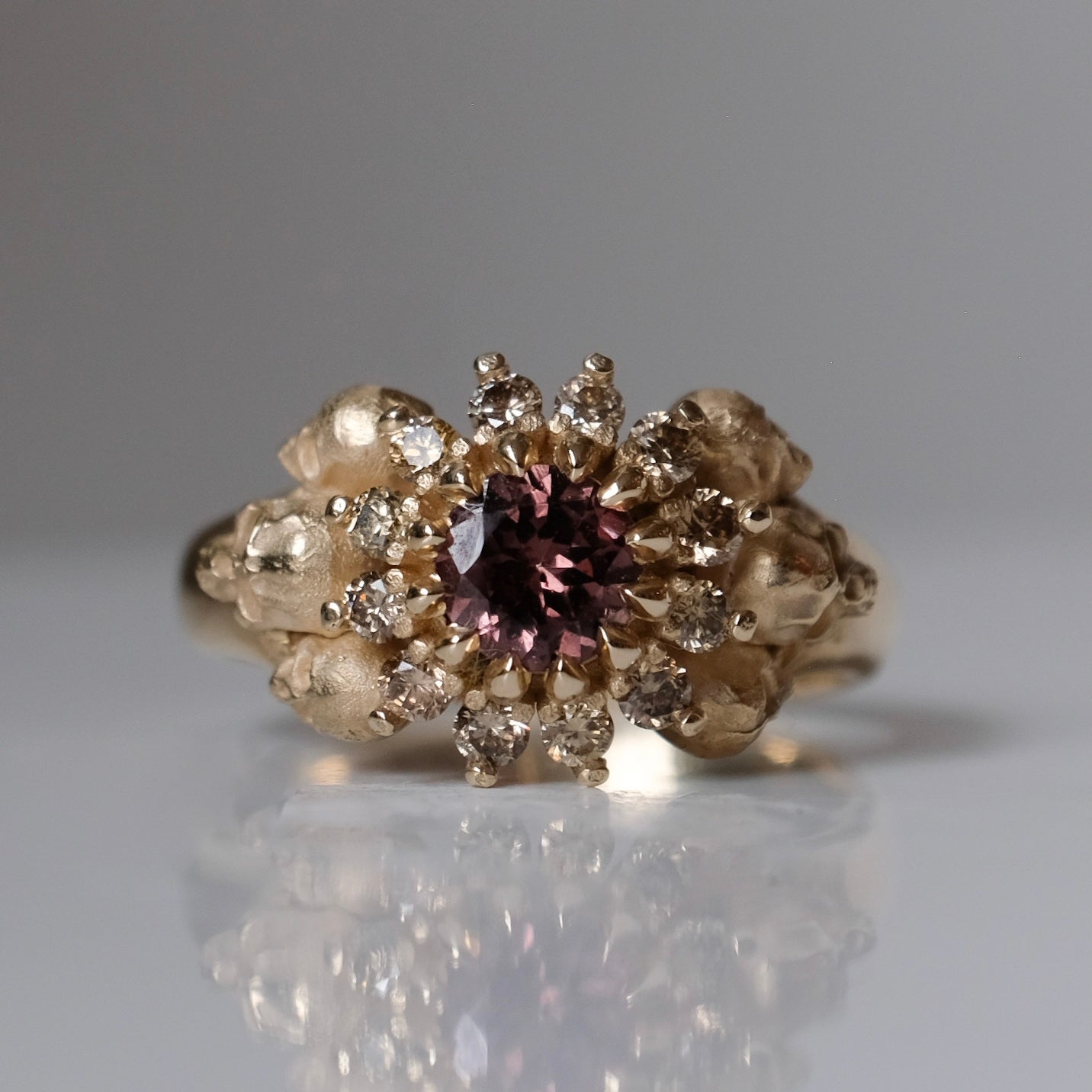 9ct yellow gold engagement ring with central colour change garnet and a champagne diamond halo. 6 skulls flank the side of the setting. 
Handmade in London by Charlie Shires