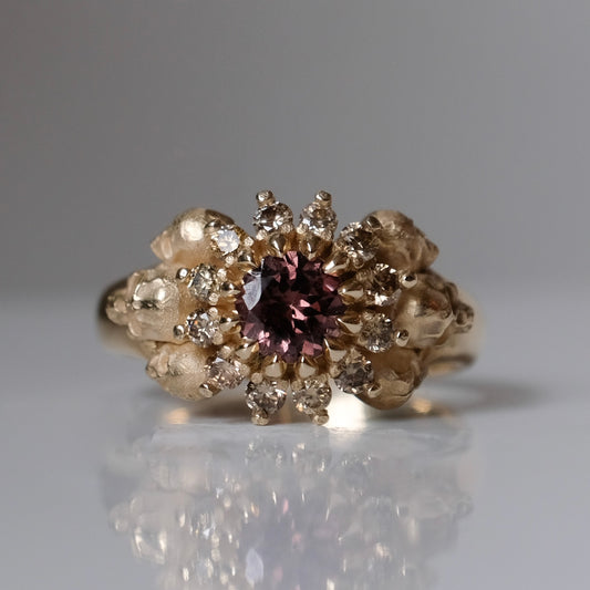 9ct yellow gold engagement ring with central colour change garnet and a champagne diamond halo. 6 skulls flank the side of the setting. 
Handmade in London by Charlie Shires