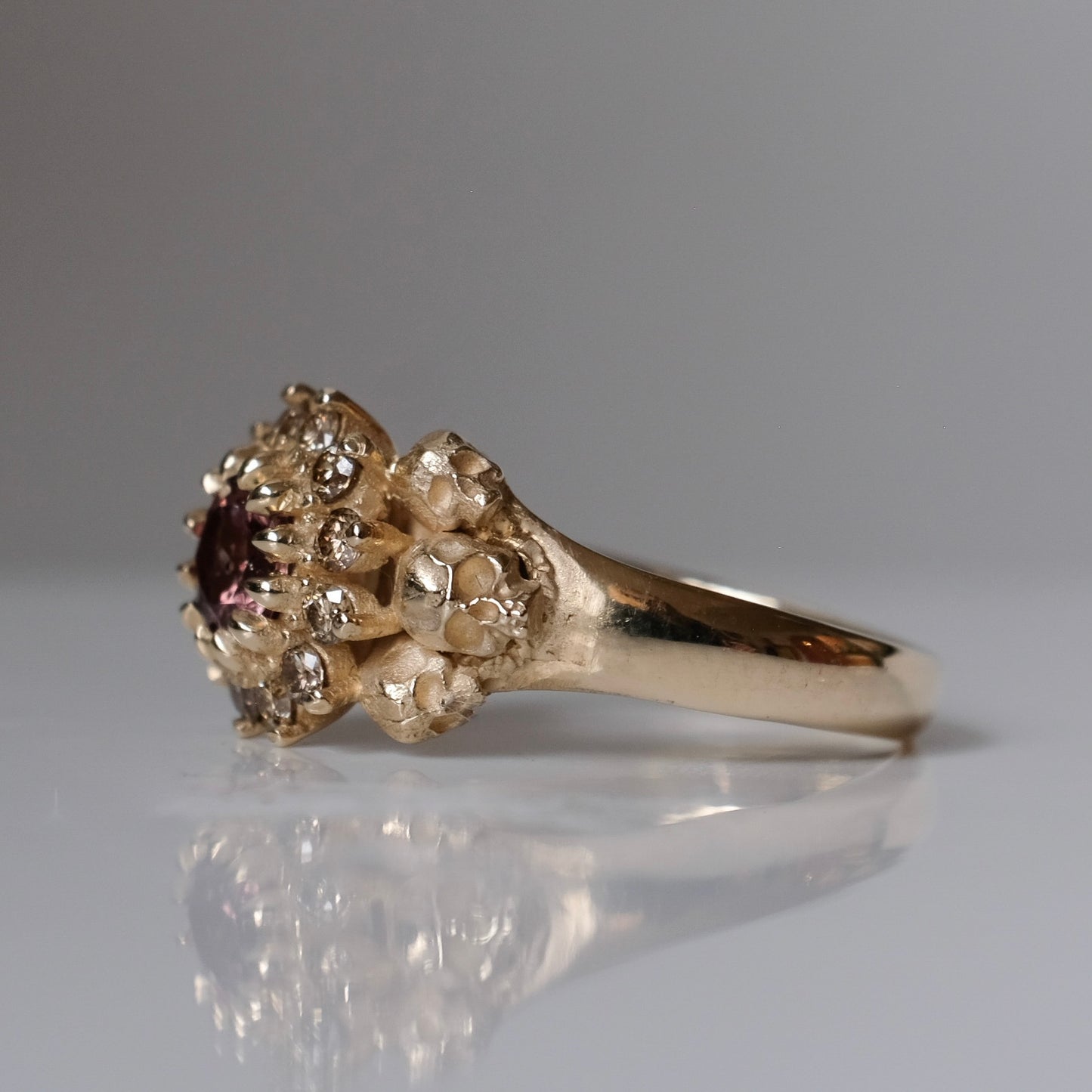 9ct yellow gold engagement ring with central colour change garnet and a champagne diamond halo. 6 skulls flank the side of the setting. 
Handmade in London by Charlie Shires