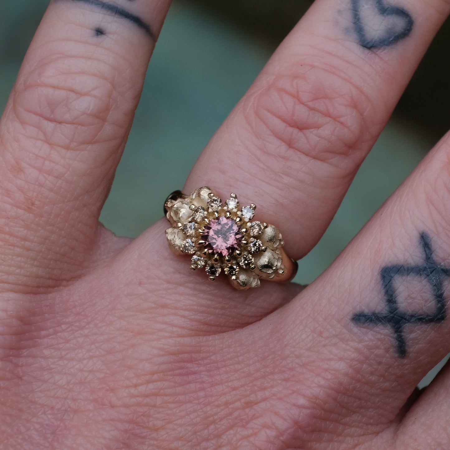 9ct yellow gold engagement ring with central colour change garnet and a champagne diamond halo. 6 skulls flank the side of the setting. 
Handmade in London by Charlie Shires