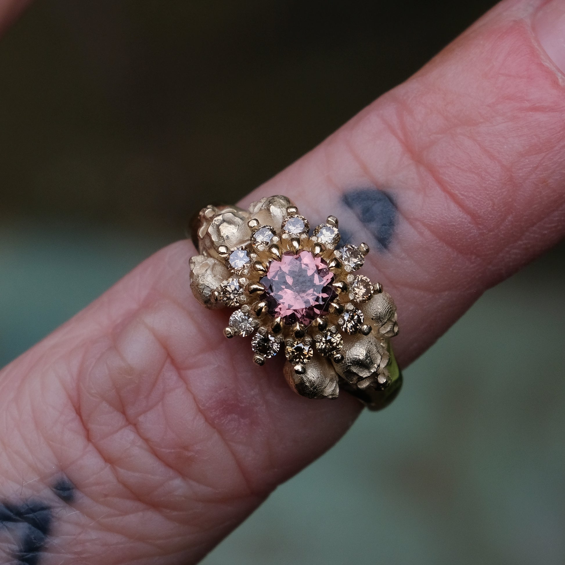 9ct yellow gold engagement ring with central colour change garnet and a champagne diamond halo. 6 skulls flank the side of the setting. 
Handmade in London by Charlie Shires