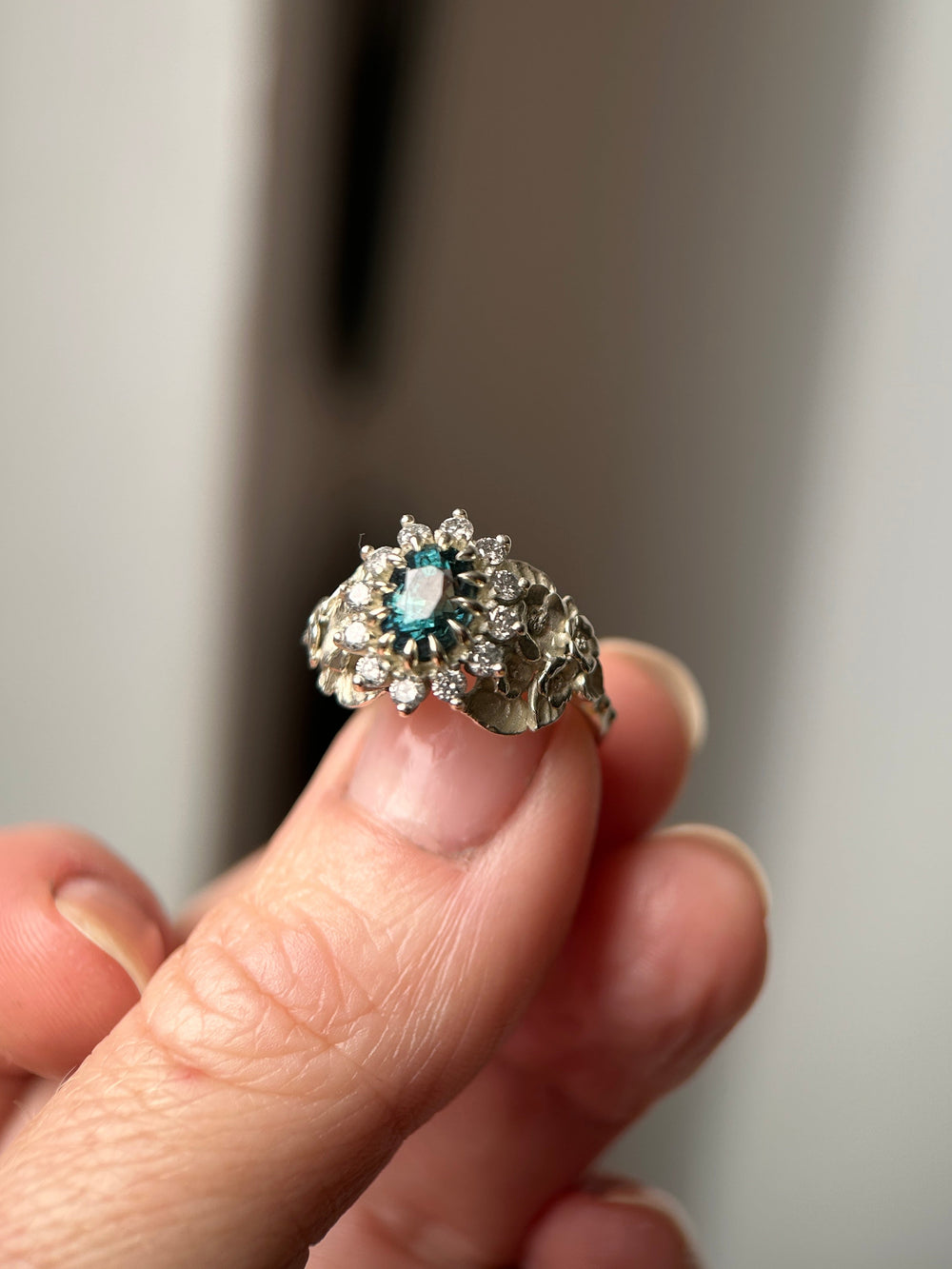 A white gold ring with a central light blue sapphire gemstone and surrounding diamonds with flowers on band, held between fingers against a soft gray background.