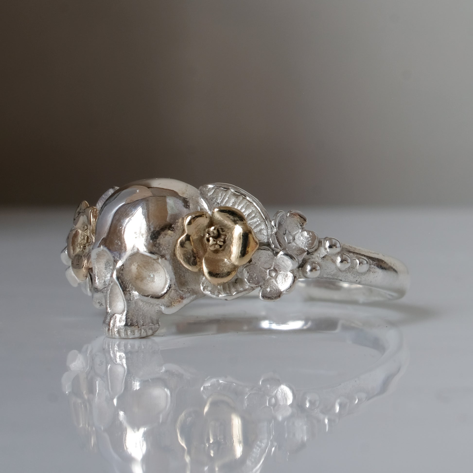 Sterling silver ring with central skull and 9ct yellow gold flower and leaves cascading down band. Handmade in London by Charlie Shires