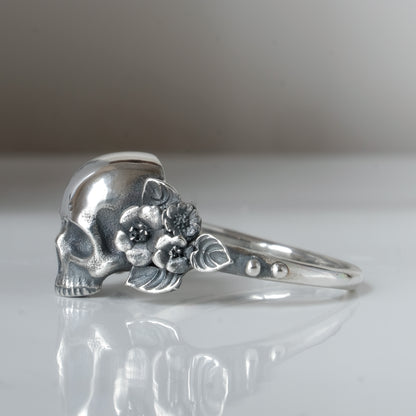 Sterling silver ring with central large skull and flower and leaves cascading down band. Handmade in London by Charlie Shires