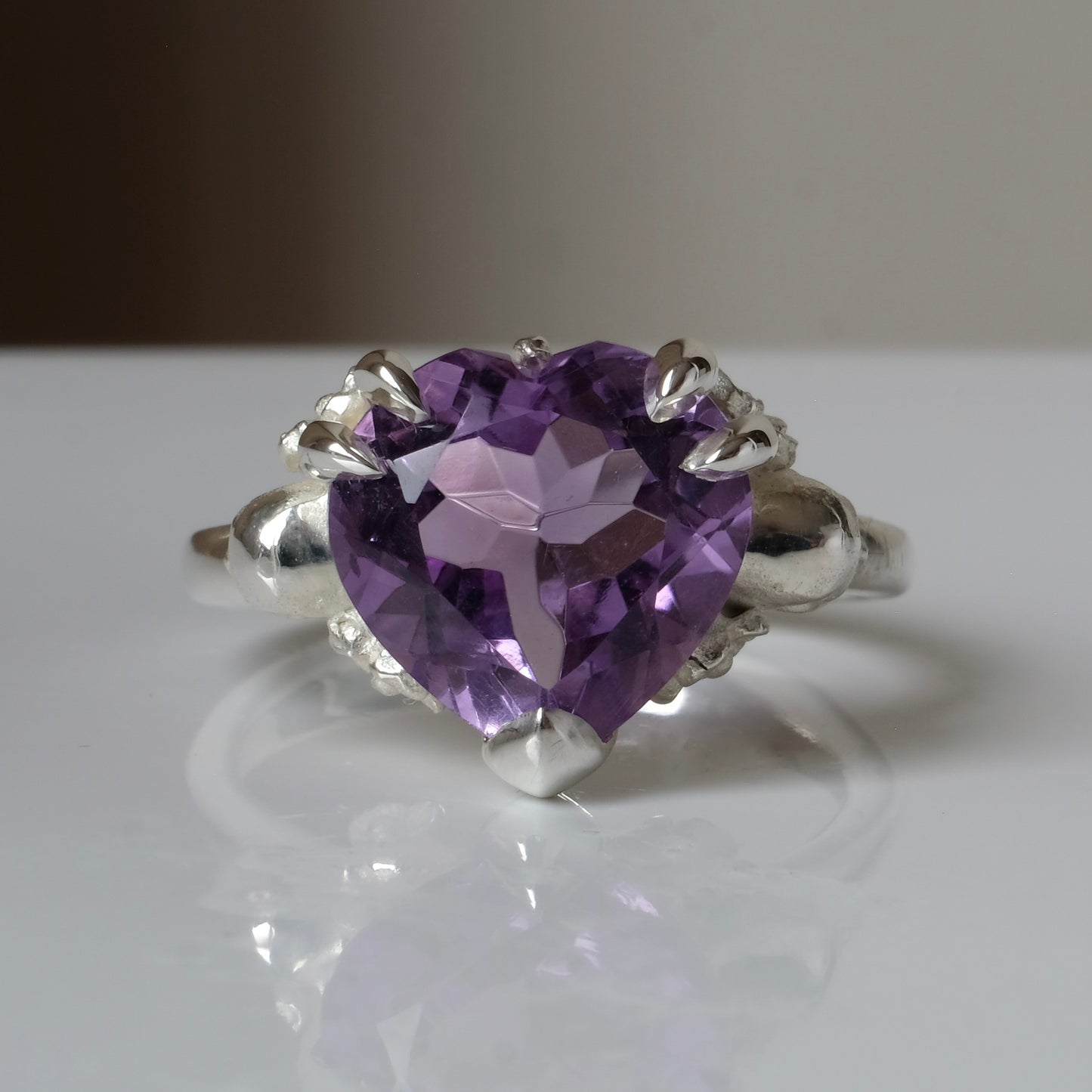 Silver ring with a heart-shaped purple amethyst gemstone decorated with skulls and flowers on a white surface