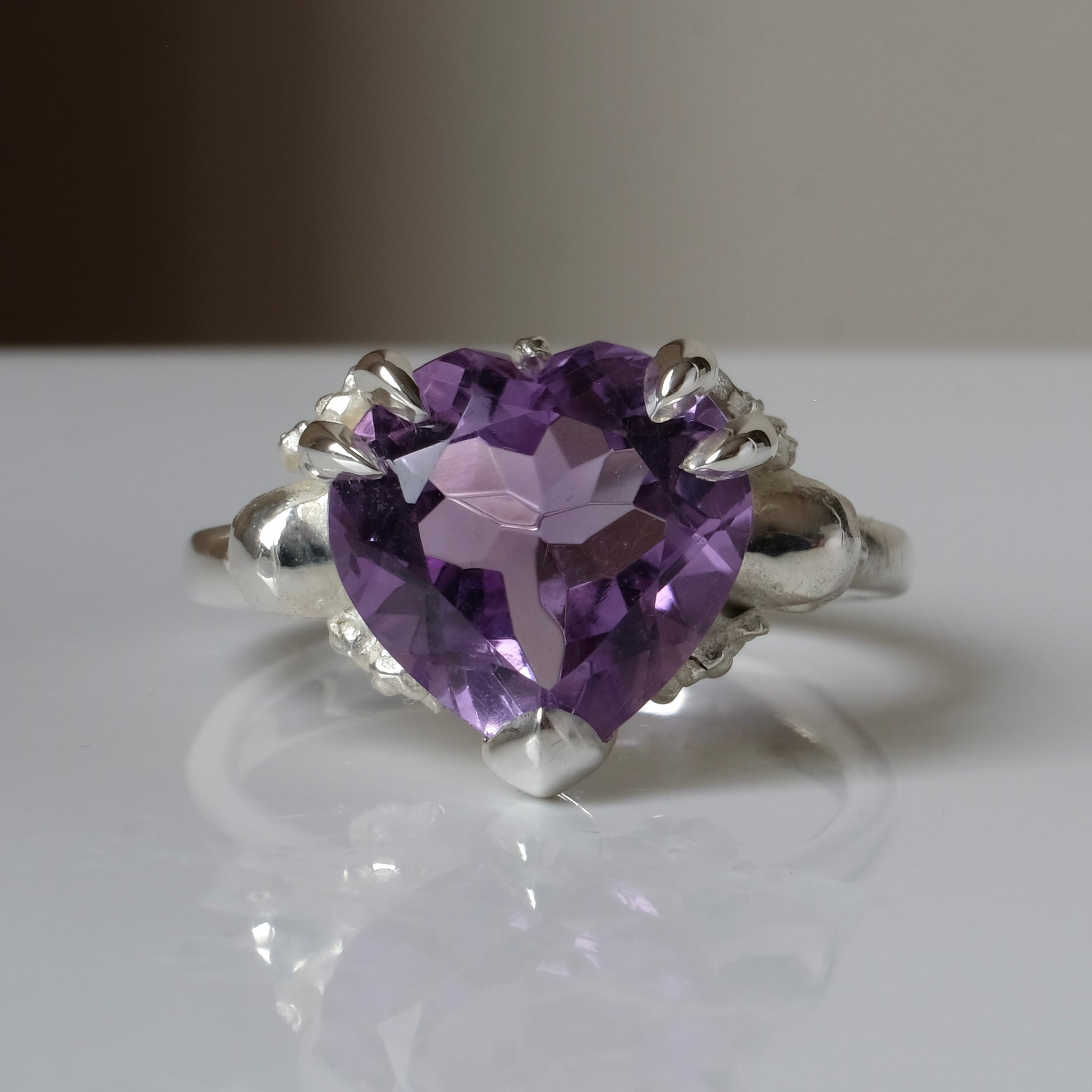 Silver ring with a heart-shaped purple amethyst gemstone decorated with skulls and flowers on a white surface