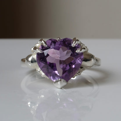 Silver ring with a heart-shaped purple amethyst gemstone decorated with skulls and flowers on a white surface