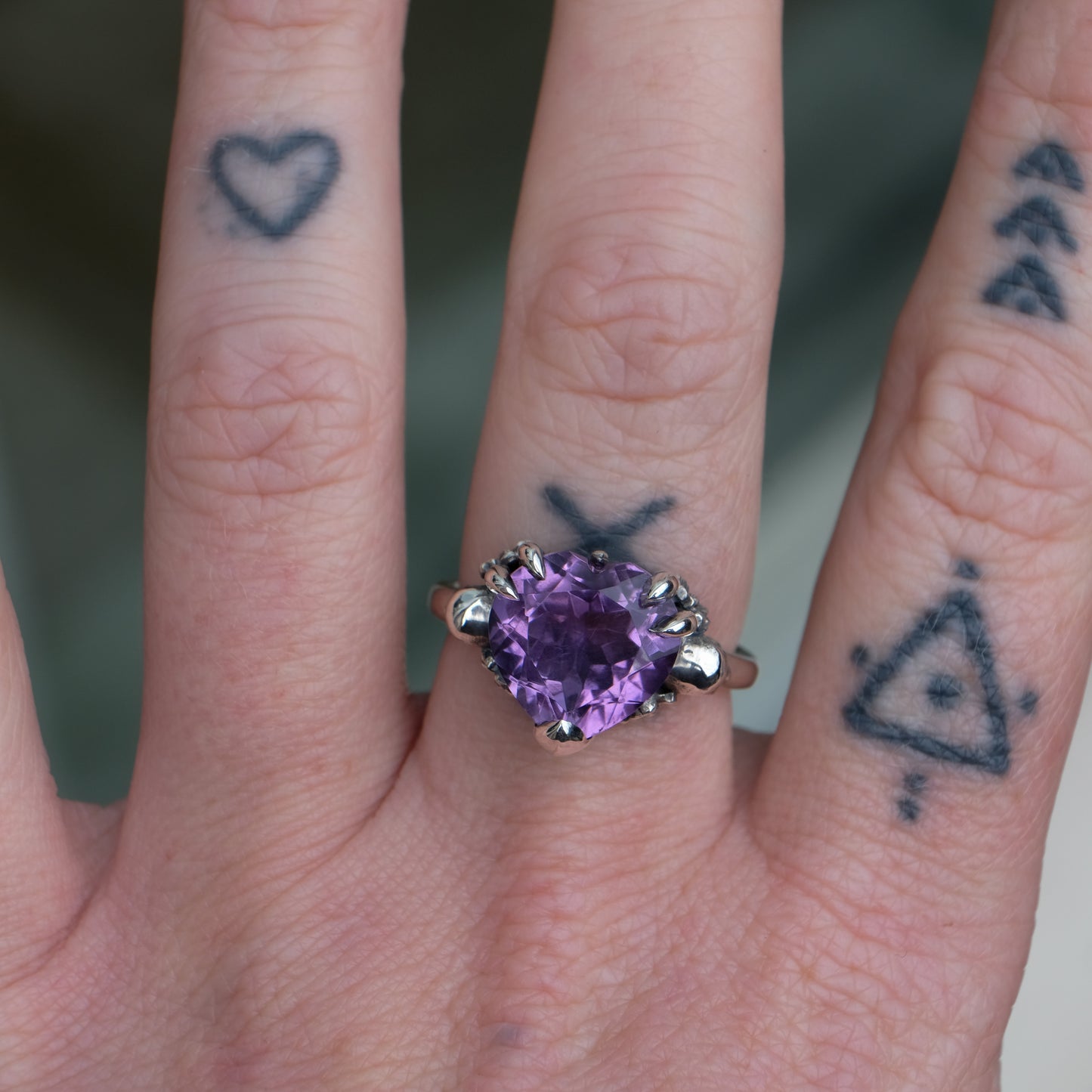 Silver ring with a heart-shaped purple amethyst gemstone decorated with skulls and flowers worn on hand with tattoos