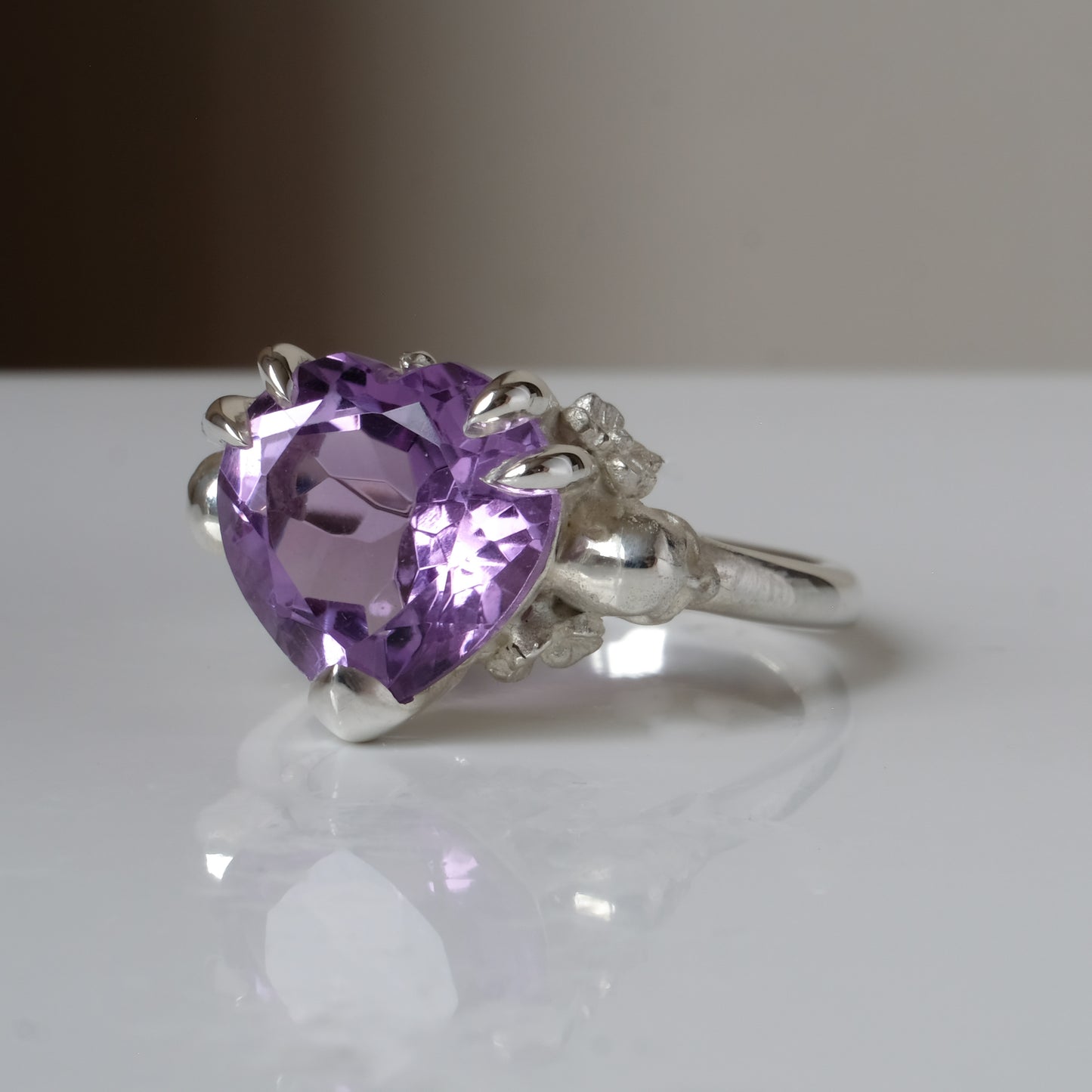 Silver ring with a heart-shaped purple amethyst gemstone decorated with skulls and flowers on a white surface
