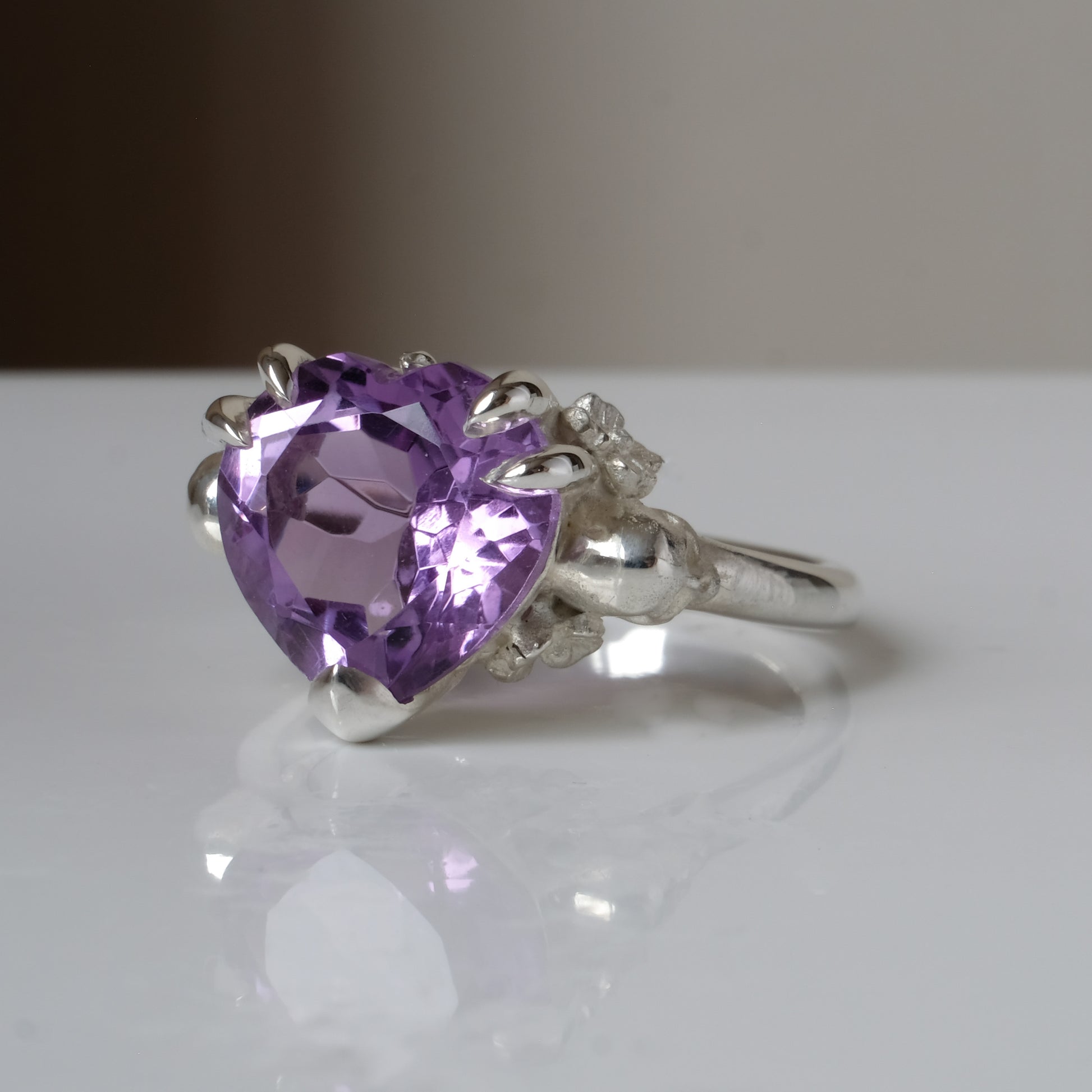 Silver ring with a heart-shaped purple amethyst gemstone decorated with skulls and flowers on a white surface