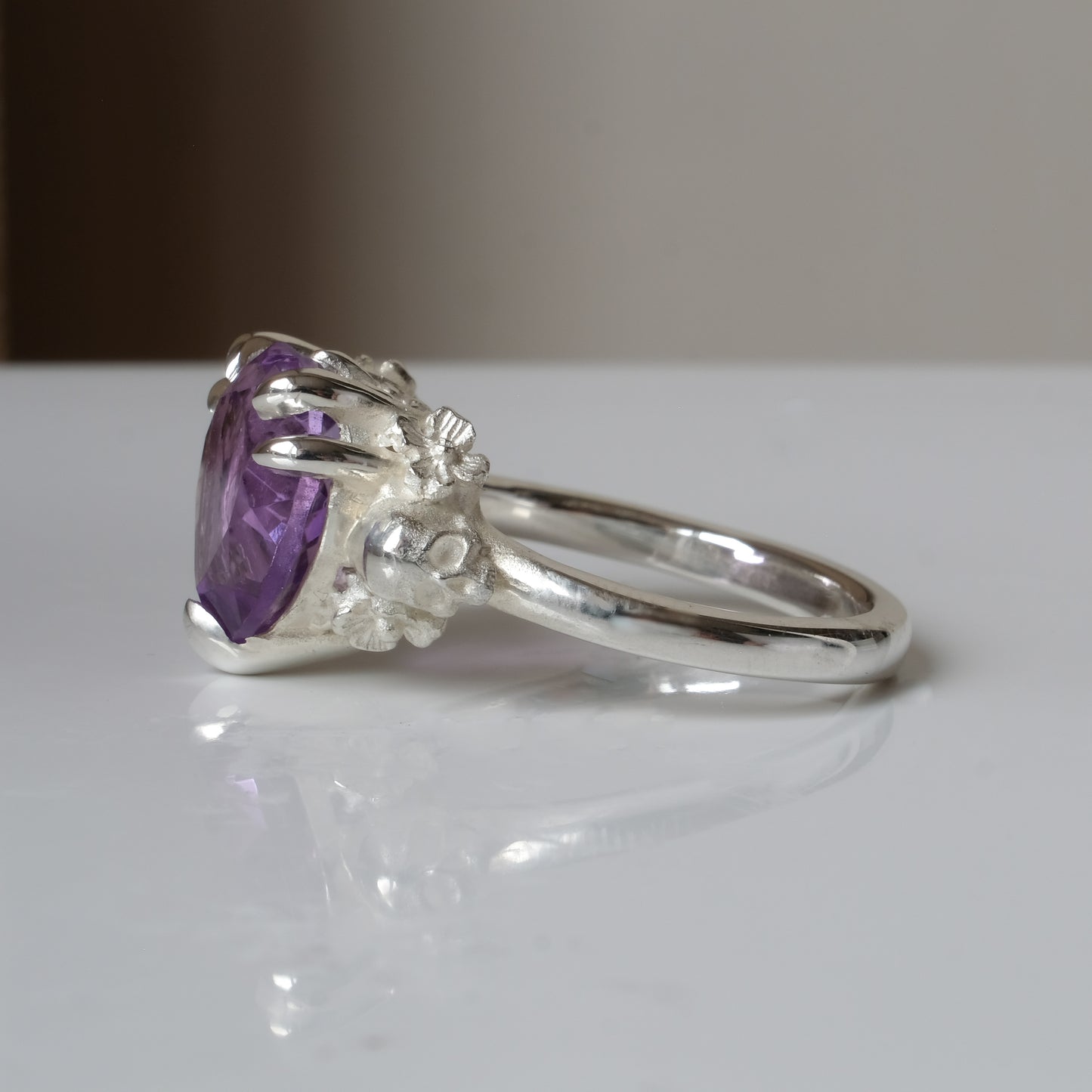 Silver ring with a heart-shaped purple amethyst gemstone decorated with skulls and flowers on a white surface