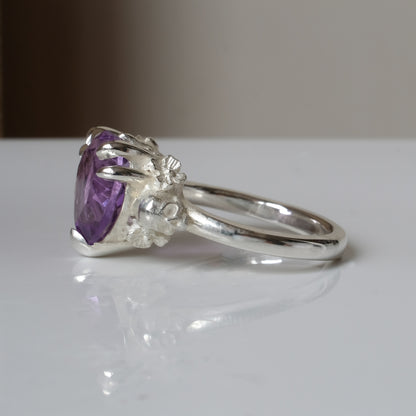 Silver ring with a heart-shaped purple amethyst gemstone decorated with skulls and flowers on a white surface