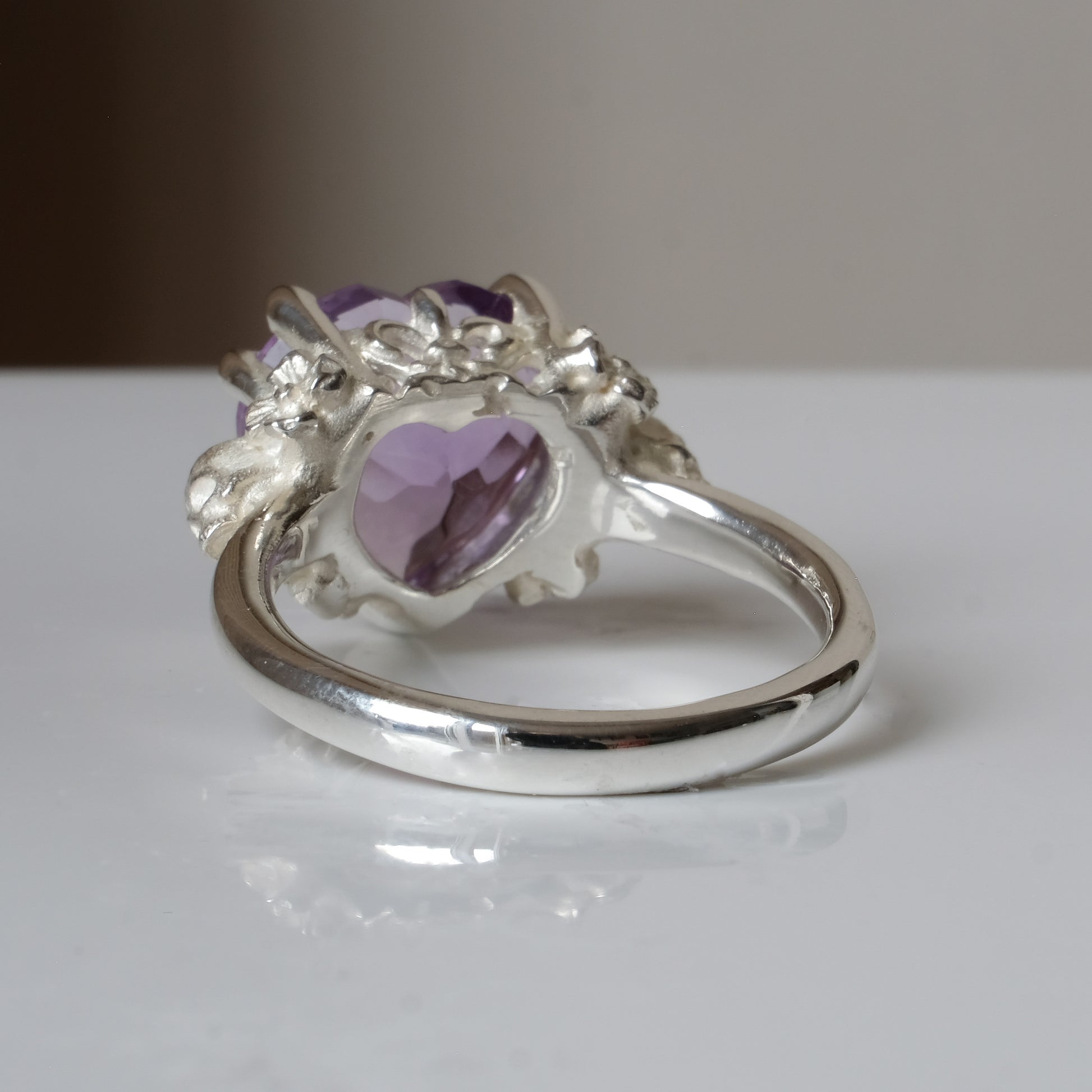 Silver ring with a heart-shaped purple amethyst gemstone decorated with skulls and flowers on a white surface