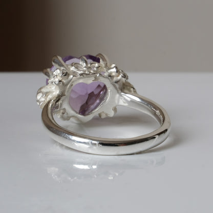 Silver ring with a heart-shaped purple amethyst gemstone decorated with skulls and flowers on a white surface