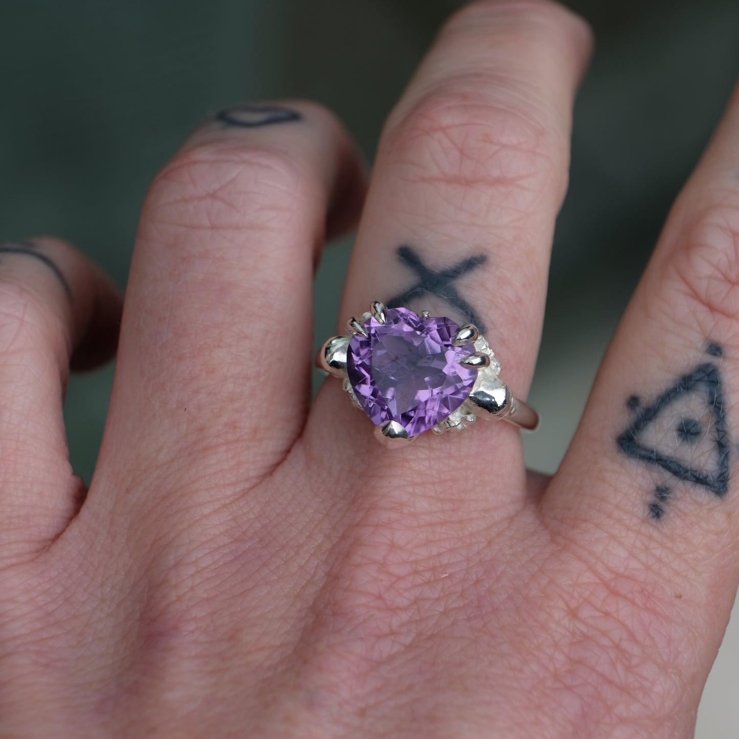 Silver ring with a heart-shaped purple amethyst gemstone decorated with skulls and flowers worn on hand with tattoos