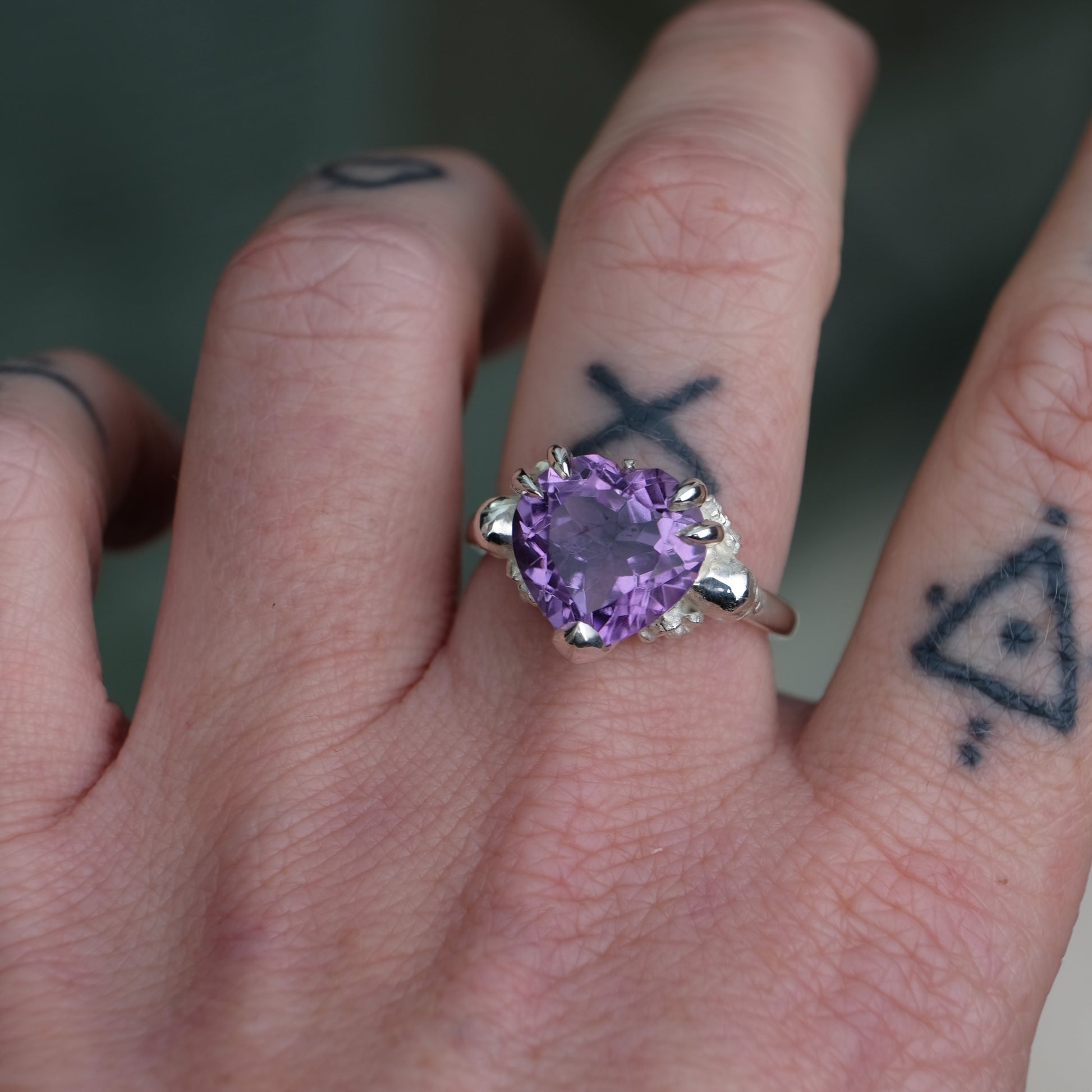 Silver ring with a heart-shaped purple amethyst gemstone decorated with skulls and flowers worn on hand with tattoos