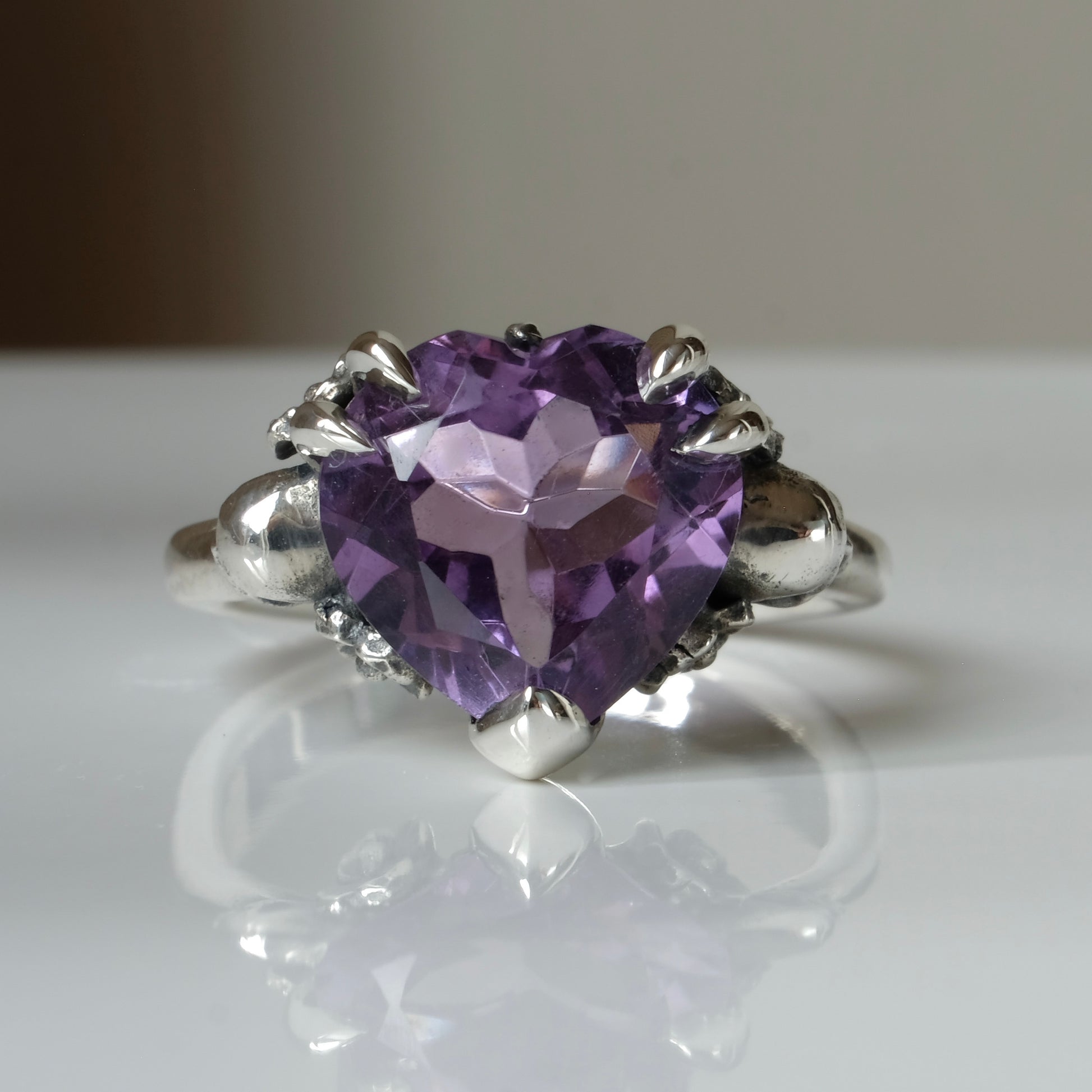 Silver ring with a heart-shaped purple amethyst gemstone decorated with skulls and flowers on a white surface
