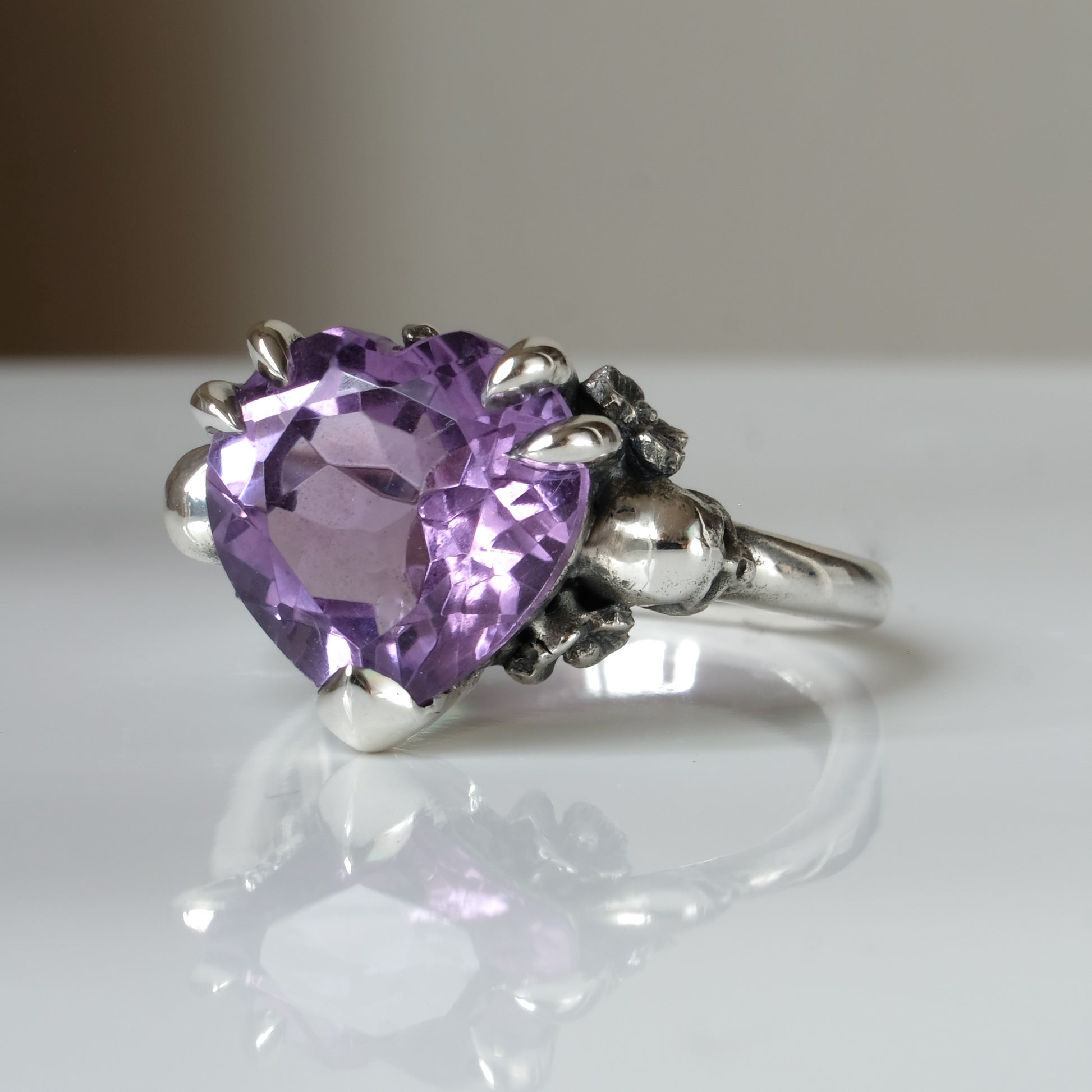 Silver ring with a heart-shaped purple amethyst gemstone decorated with skulls and flowers on a white surface