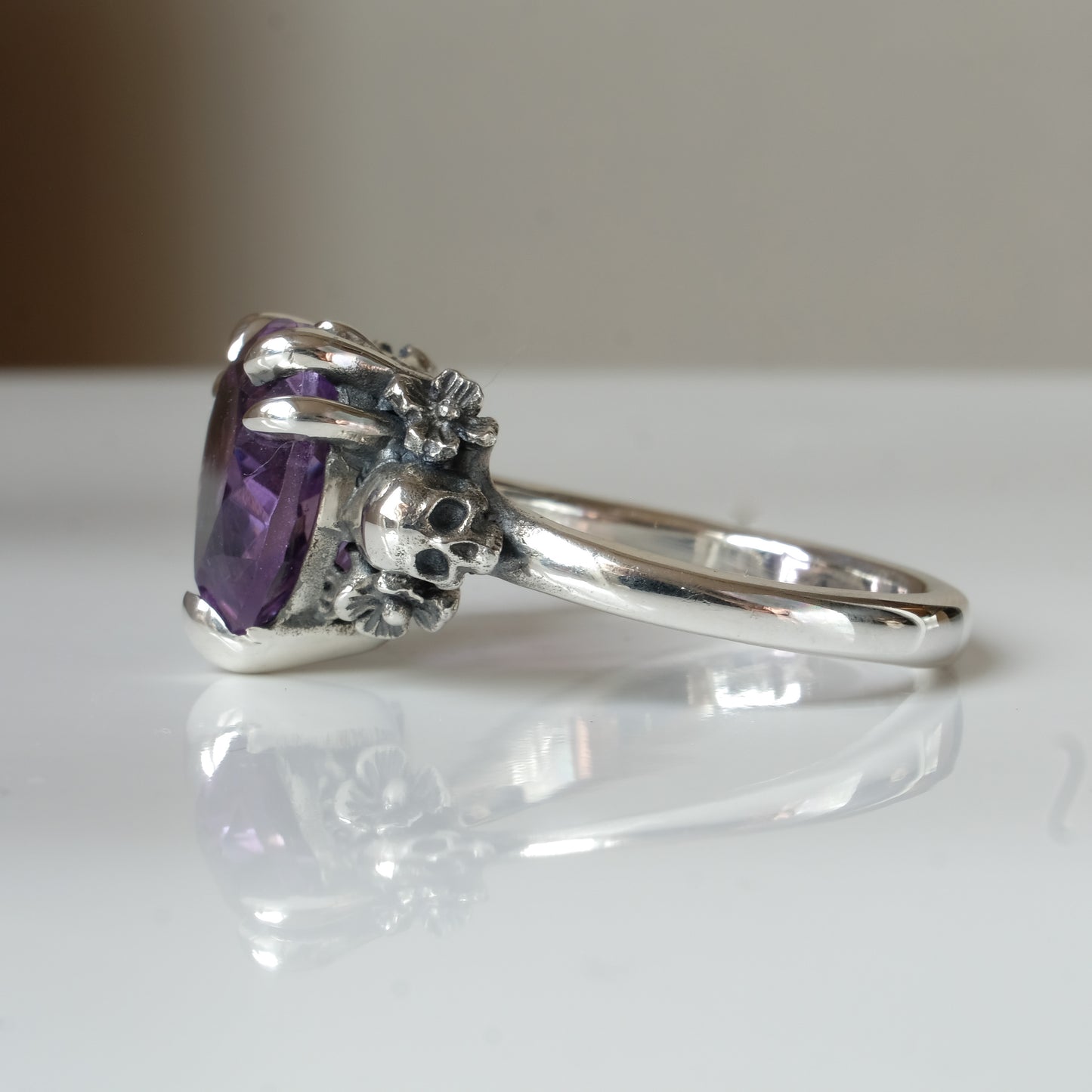 Silver ring with a heart-shaped purple amethyst gemstone decorated with skulls and flowers on a white surface