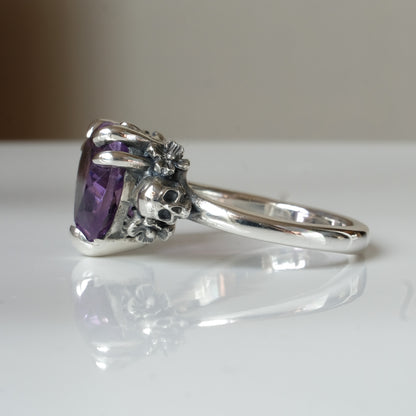 Silver ring with a heart-shaped purple amethyst gemstone decorated with skulls and flowers on a white surface