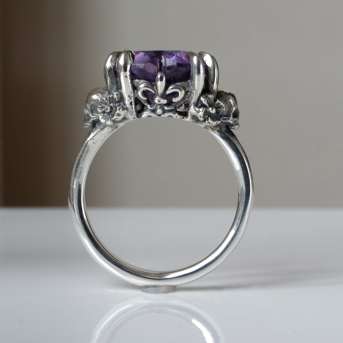 Silver ring with a heart-shaped purple amethyst gemstone decorated with skulls and flowers on a white surface