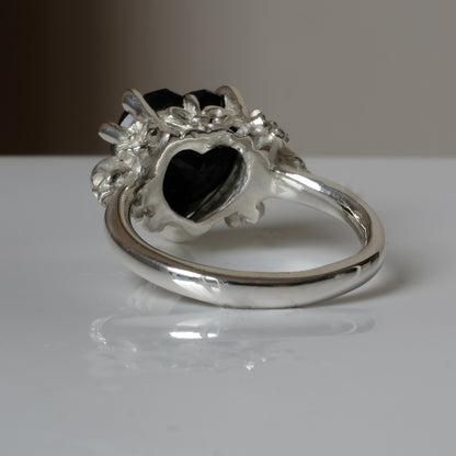 Silver ring with a heart-shaped black spinel gemstone decorated with skulls and flowers on a white surface