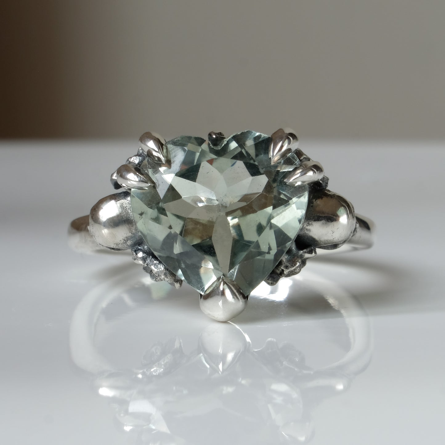Silver ring with a heart-shaped green mint quartz gemstone decorated with skulls and flowers on a white surface