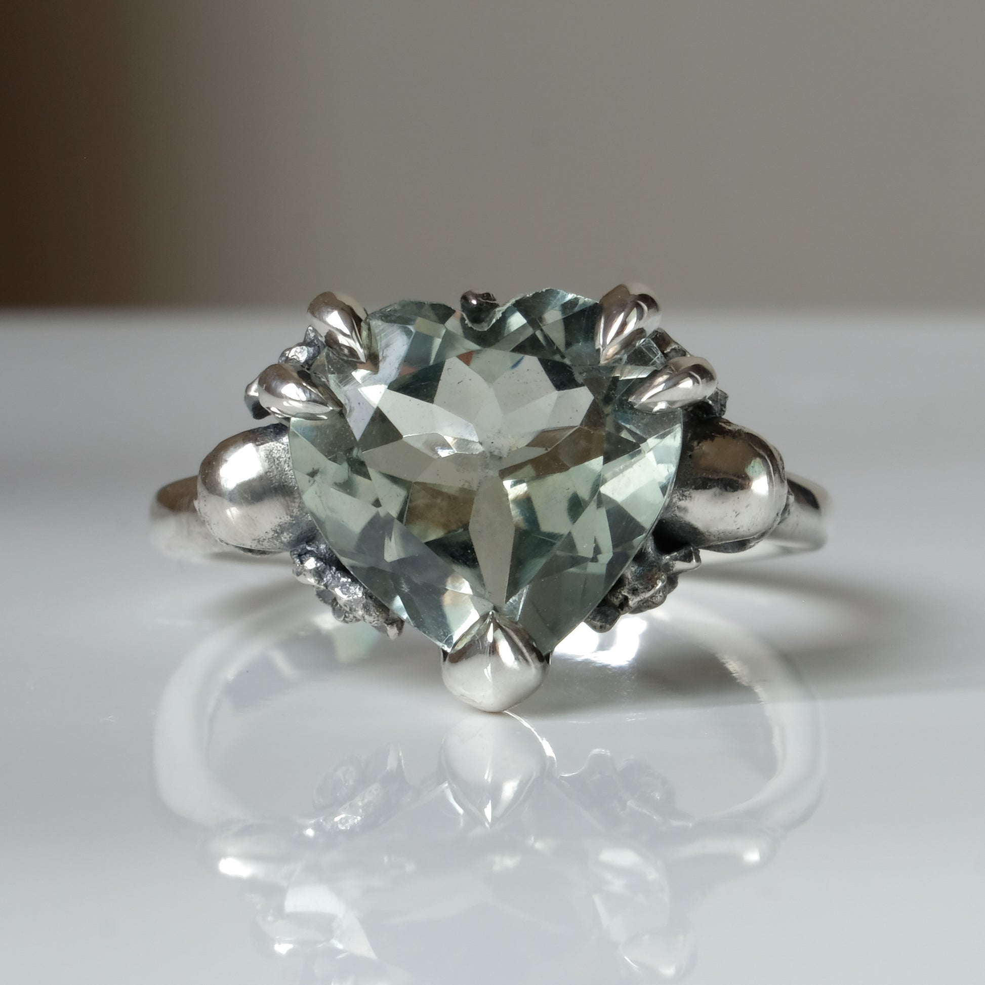 Silver ring with a heart-shaped green mint quartz gemstone decorated with skulls and flowers on a white surface