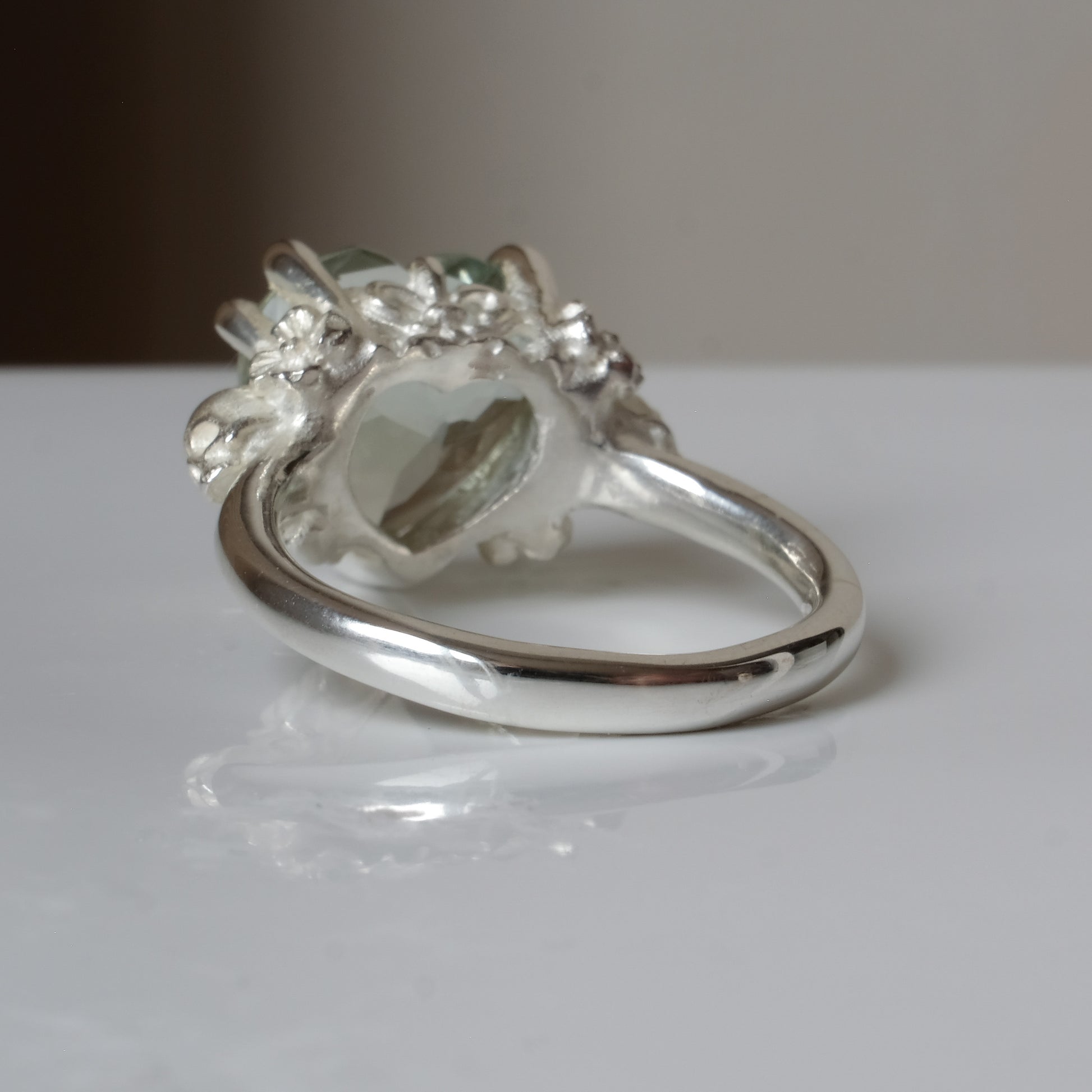 Silver ring with a heart-shaped green mint quartz gemstone decorated with skulls and flowers on a white surface