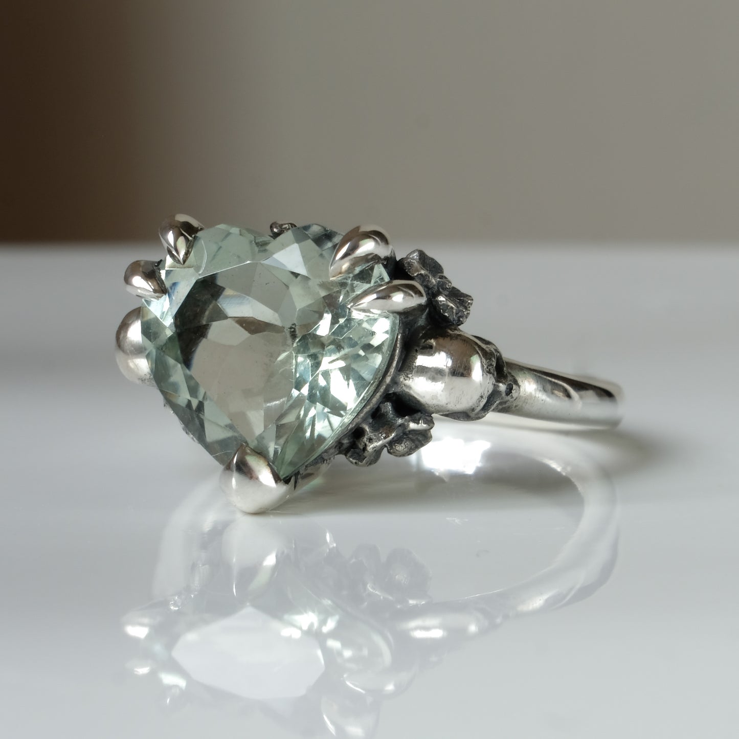 Silver ring with a heart-shaped green mint quartz gemstone decorated with skulls and flowers on a white surface
