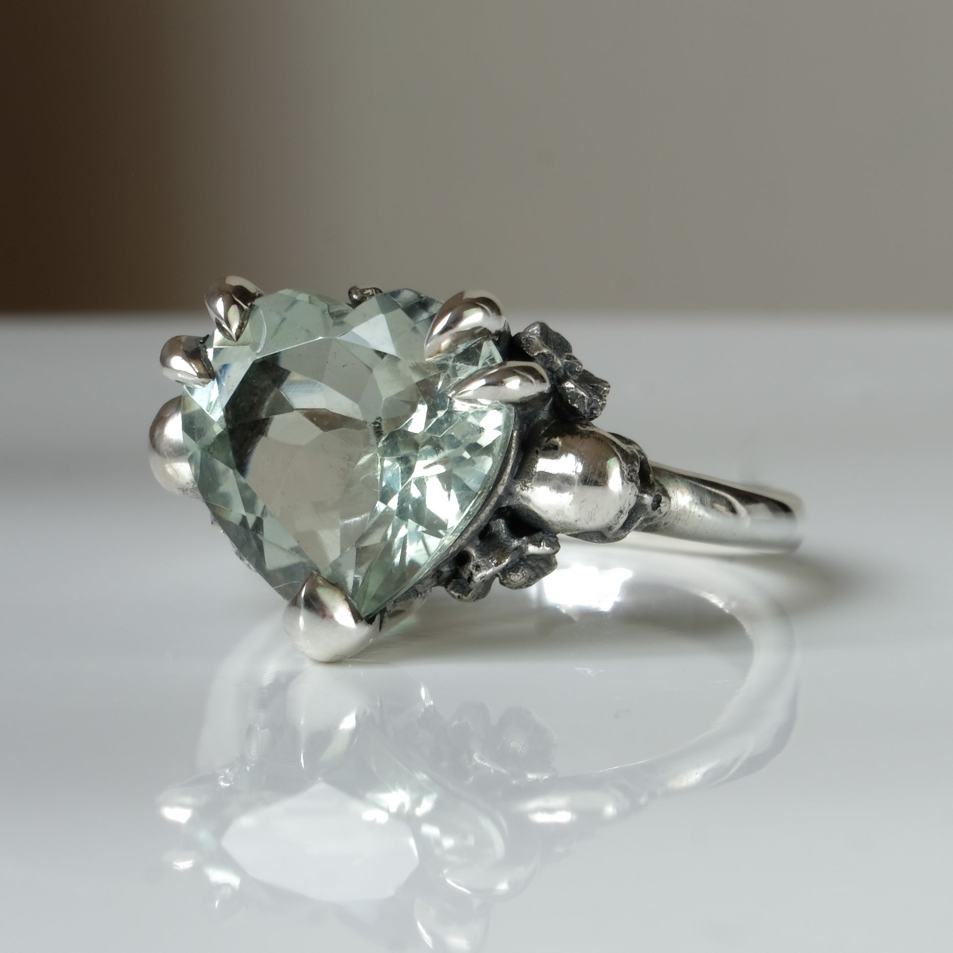 Silver ring with a heart-shaped green mint quartz gemstone decorated with skulls and flowers on a white surface