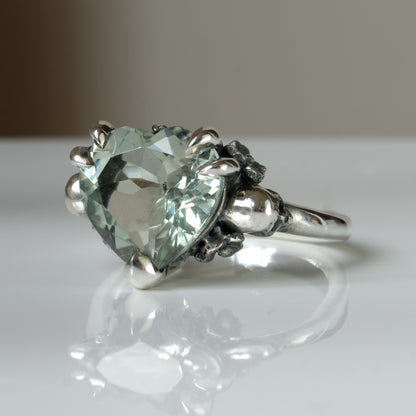 Silver ring with a heart-shaped green mint quartz gemstone decorated with skulls and flowers on a white surface
