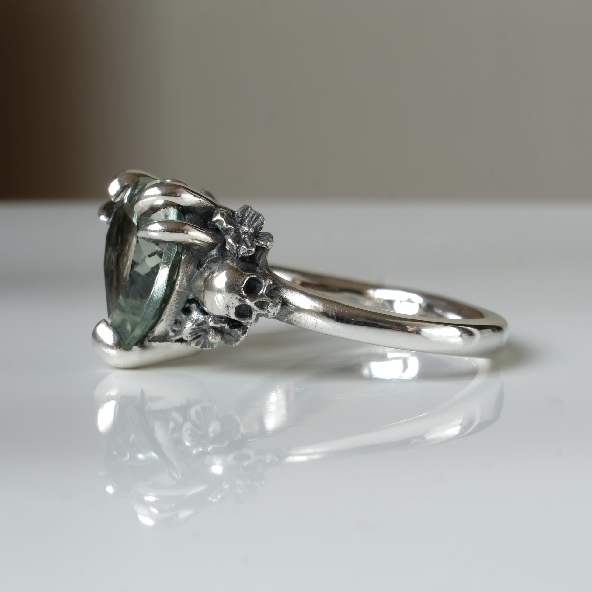 Silver ring with a heart-shaped green mint quartz gemstone decorated with skulls and flowers on a white surface