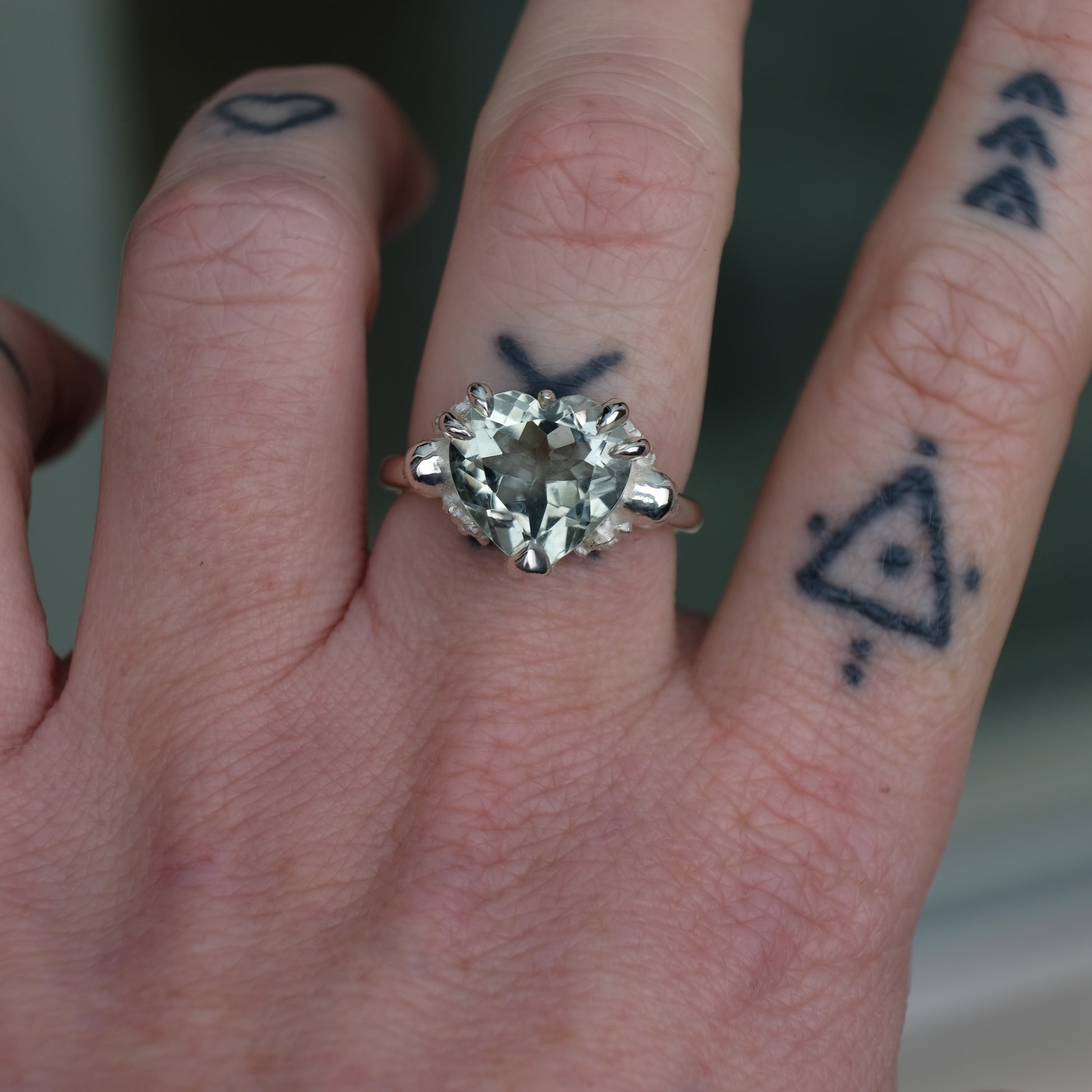 Silver ring with a heart-shaped green mint quartz gemstone decorated with skulls and flowers worn on hand with tattoos