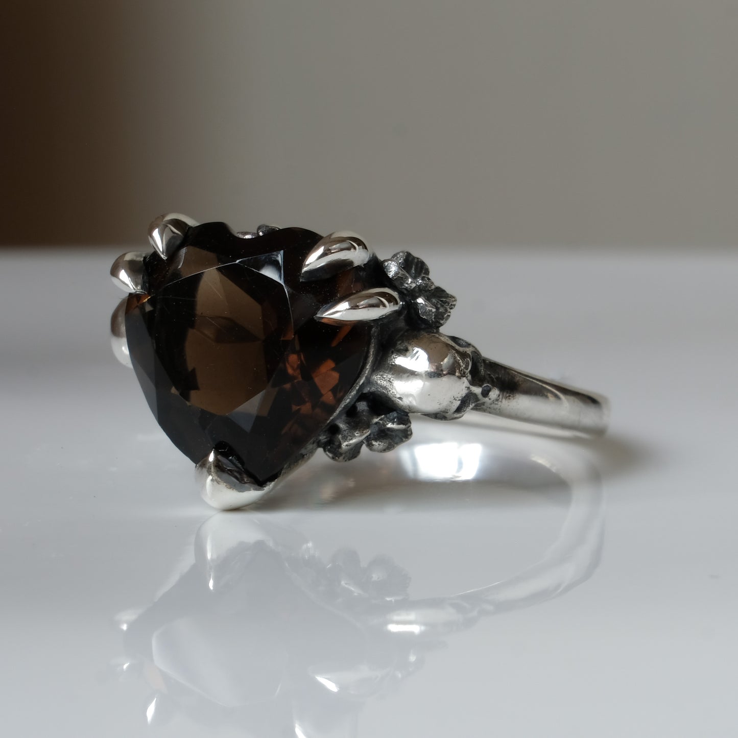 Silver ring with a heart-shaped brown smokey quartz gemstone decorated with skulls and flowers on a white surface