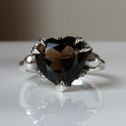 Silver ring with a heart-shaped brown smokey quartz gemstone decorated with skulls and flowers on a white surface