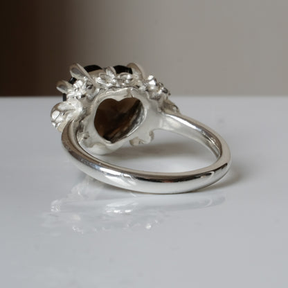 Silver ring with a heart-shaped brown smokey quartz gemstone decorated with skulls and flowers on a white surface