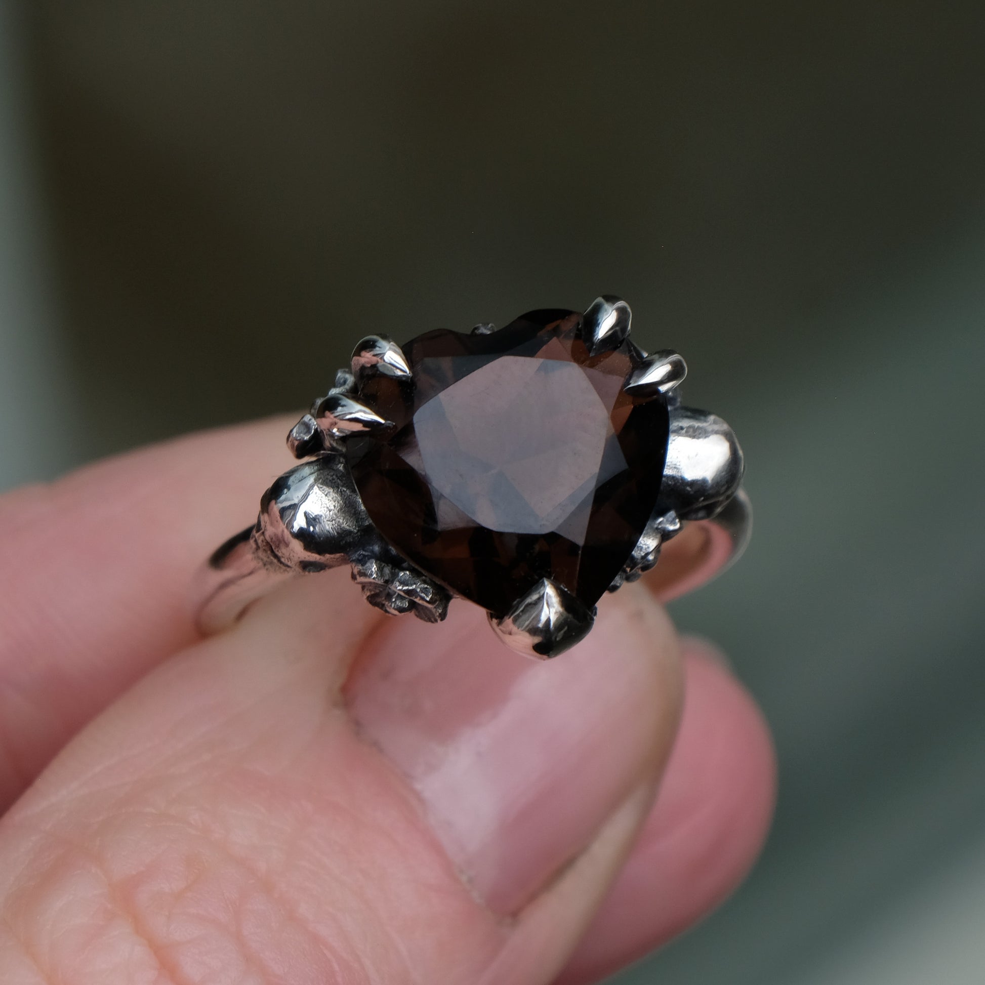 Silver ring with a heart-shaped brown smokey quartz gemstone decorated with skulls and flowers upclose held by fingers