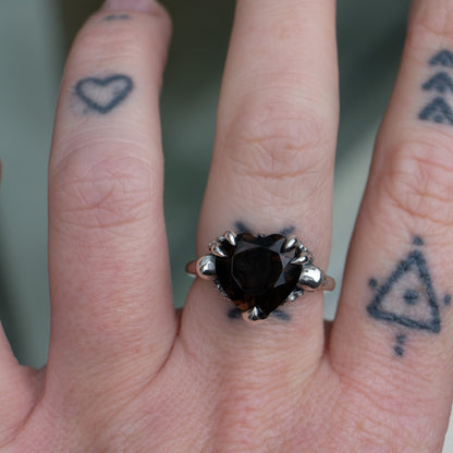 Silver ring with a heart-shaped brown smokey quartz gemstone decorated with skulls and flowers worn on hand with tattoos