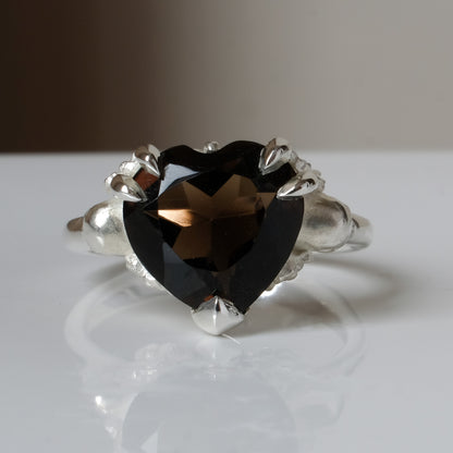 Silver ring with a heart-shaped brown smokey quartz gemstone decorated with skulls and flowers on a white surface
