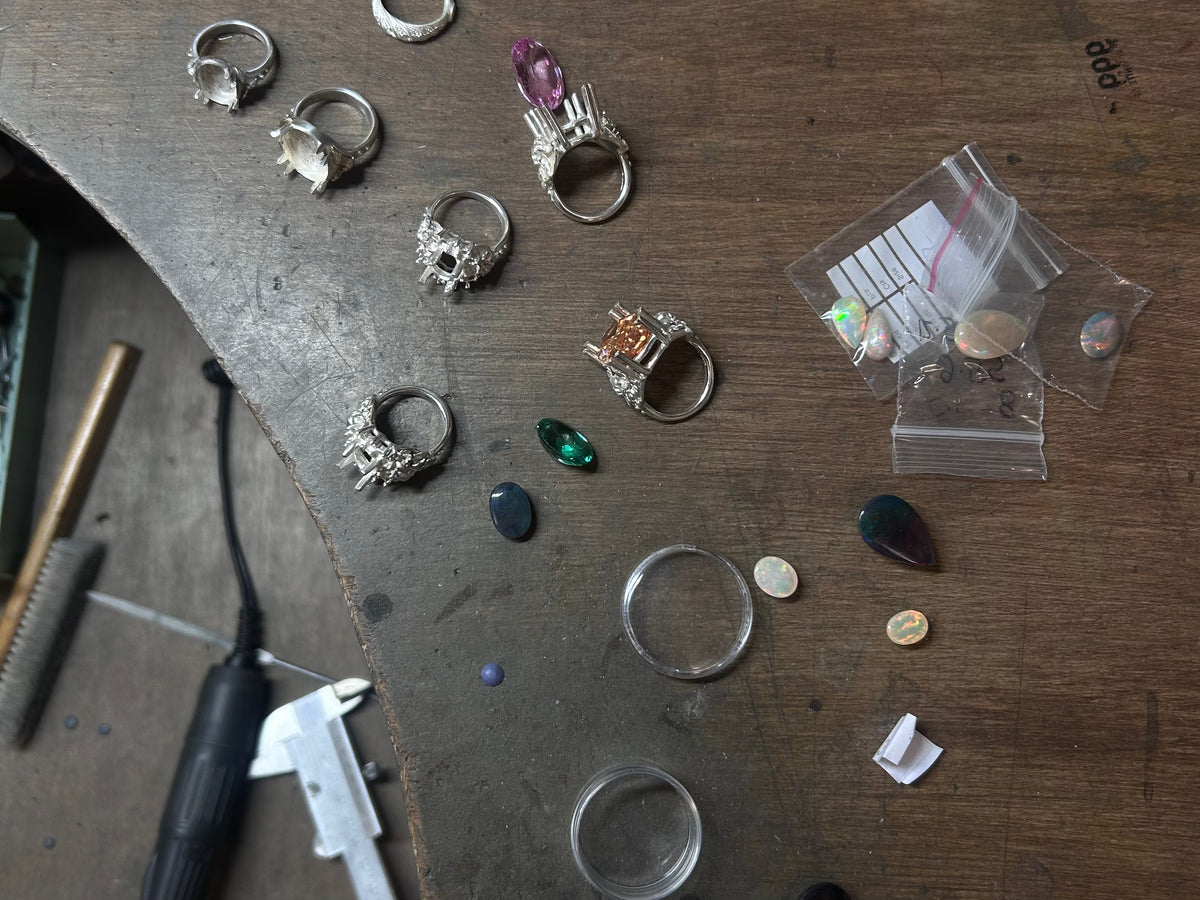 A collection of handmade silver rings with various gemstones and decorative settings, displayed on a wooden workbench alongside tools by Charlie Shires