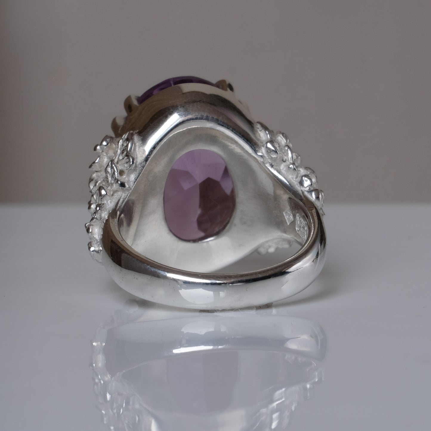 Ring claw set with large amethyst gemstone with skull and floral detail on side.  made in sterling silver on reflective surface