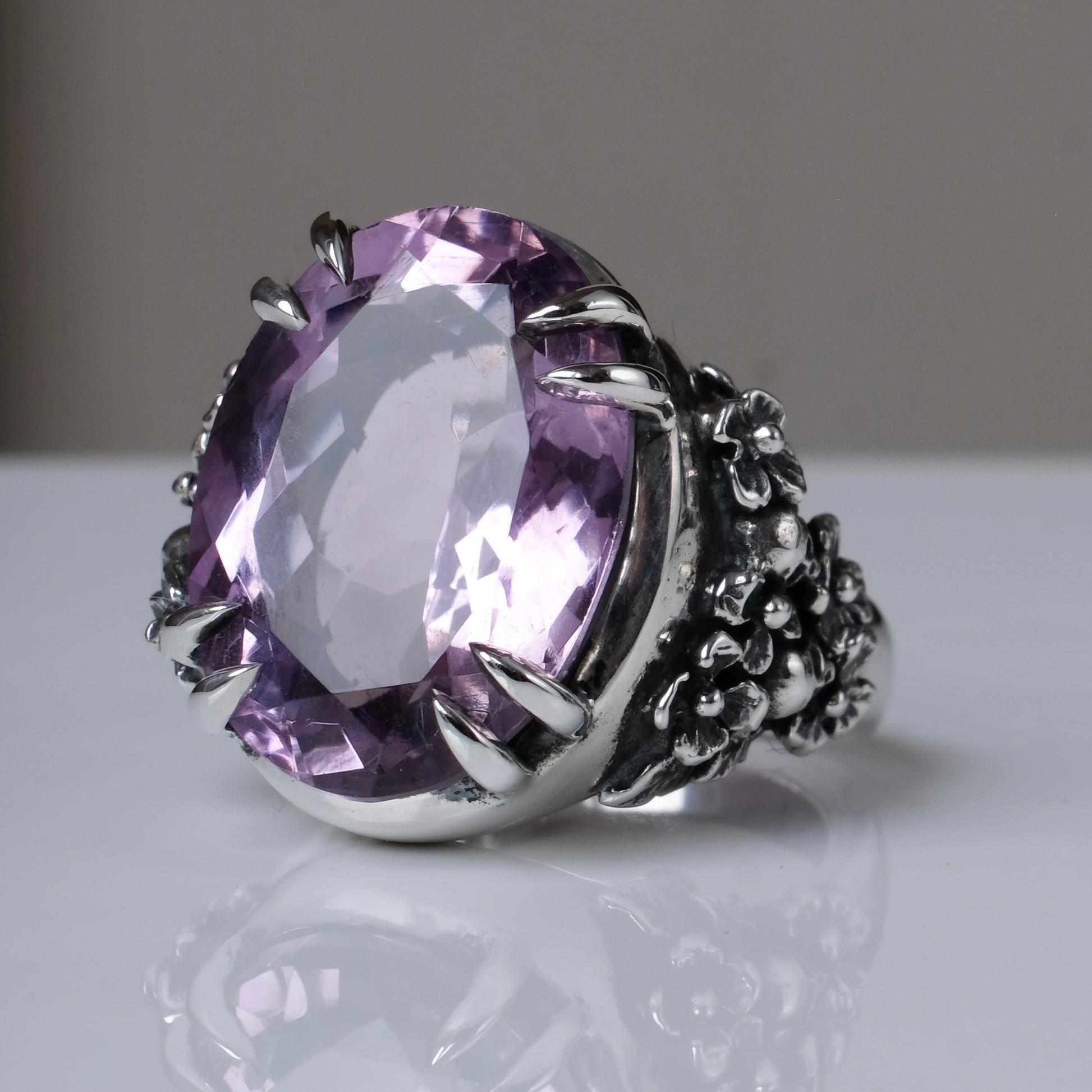 Ring claw set with large amethyst gemstone with skull and floral detail on side., made in sterling silver on white background