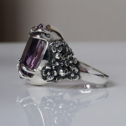 Ring claw set with large amethyst gemstone with skull and floral detail on side.  made in sterling silver on reflective surface