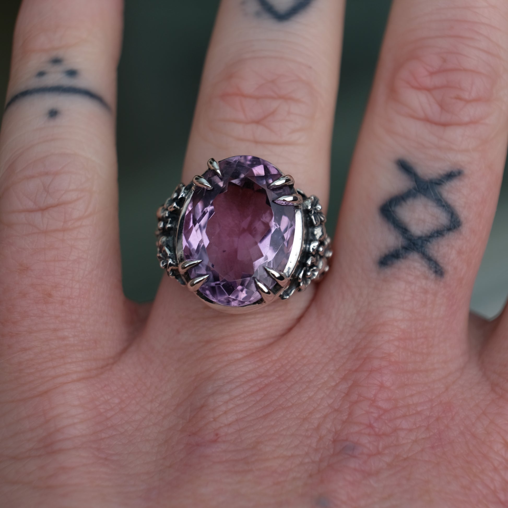 Ring claw set with large amethyst gemstone with skull and floral detail on side.  made in sterling silver worn on hand with tattoos