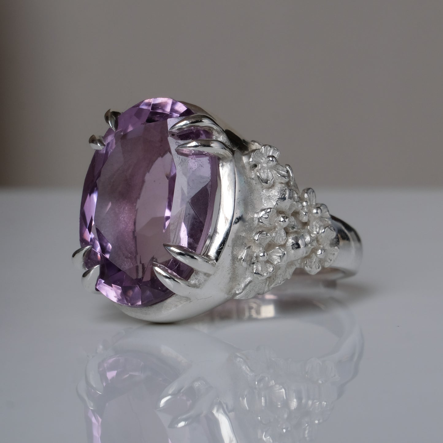 Ring claw set with large amethyst gemstone with skull and floral detail on side.  made in sterling silver on reflective surface